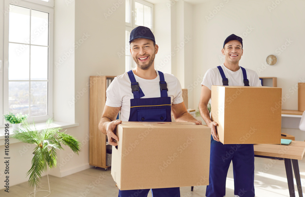 Moving servise. Two cheerful male porters from moving service carry cardboard boxes with things. Portrait of smiling professional movers in overalls help client move out of apartment or office. Moving servise. Two cheerful male porters from moving service carry cardboard boxes with things. Portrait of smiling professional movers in overalls help client move out of apartment or office.