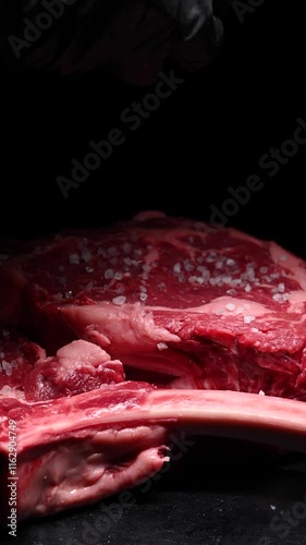 A close-up shot of marbled beef sprinkled with salt. A large piece of red meat on the bone lying on a black kitchen board.