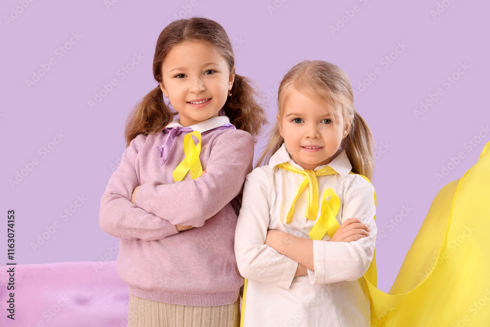 Cute little girls in superhero costume with yellow ribbons on lilac background. Childhood cancer awareness concept Cute little girls in superhero costume with yellow ribbons on lilac background. Childhood cancer awareness concept