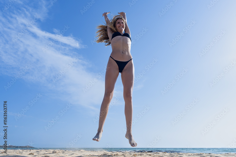 Naklejka premium Mujer joven saltando de emoción porque esta feliz y divertida, con un sensacional día de vacaciones en verano en la playa, soleado con cielo azul.