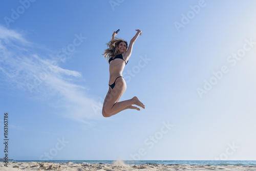 Mujer joven saltando de emoción porque esta feliz y divertida, con un sensacional día de vacaciones en verano en la playa, soleado con cielo azul. photo