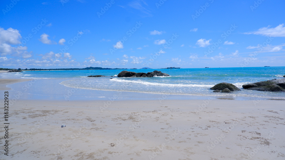 Fototapeta premium A pristine tropical beach with clear turquoise water, soft white sand, and large granite rocks. The sky is clear blue with scattered white clouds.