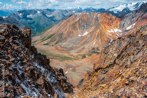 Top view from abyss edge between rocks to multicolor valley with iron river and big sharp rocky ridge of red color. Colorful large mountains in freshly fallen snow in low clouds. Vivid alpine scenery. photo