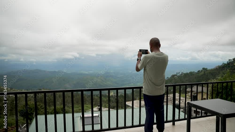A man photographs a picturesque valley scenery using a smartphone while standing on a balcony. A man photographs a picturesque valley scenery using a smartphone while standing on a balcony.
