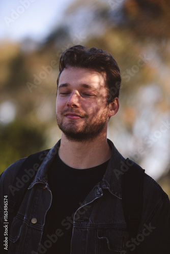 A young man with short, wavy hair stands outdoors under soft sunlight, his face partially shadowed by leaves. He's wearing a dark jacket and backpack, with blurred trees in the background