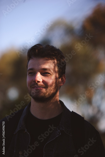 A young man with short, wavy hair stands outdoors under soft sunlight, his face partially shadowed by leaves. He's wearing a dark jacket and backpack, with blurred trees in the background