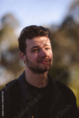 A young man with short, wavy hair stands outdoors under soft sunlight, his face partially shadowed by leaves. He's wearing a dark jacket and backpack, with blurred trees in the background