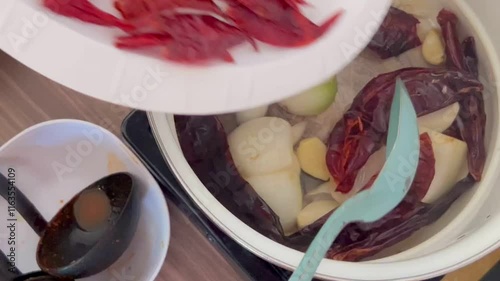 Hand stirs a mixture of dried chili and chopped onions, fresh ingredients in a pot. The setting reflects a home kitchen where traditional cooking techniques are being highlighted. photo