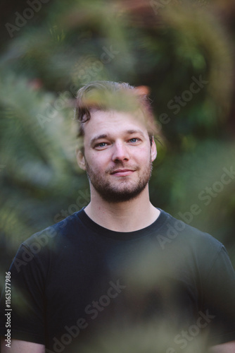 A handsome man in a black shirt stands surrounded by lush greenery, bathed in soft natural light. The background blurs with vibrant foliage, creating a peaceful and serene atmosphere