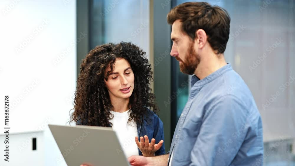 Two colleagues collaborate on a project, utilizing a laptop in a well-lit modern office. Professional woman presenting information to a bearded businessman.