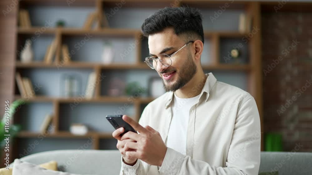 Handsome man using mobile phone sitting on sofa in living room at home. Smiling male browses social networks, does shopping in an online store, chats with a friend, writes or reads messages. Close up Handsome man using mobile phone sitting on sofa in living room at home. Smiling male browses social networks, does shopping in an online store, chats with a friend, writes or reads messages. Close up