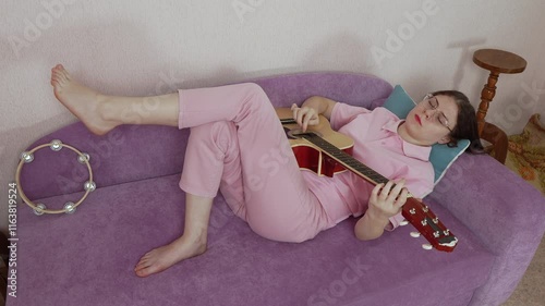 Woman guitarist with eyeglasses and her eyes closed laying on her back on sofa and playing guitar. Musician of Caucasian ethnicity dressed casually in jeans, polo shirt. High angle view, handheld shot