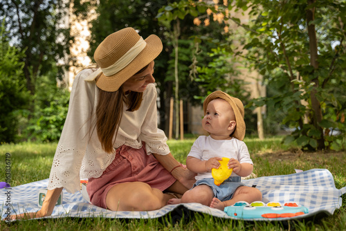 Mother and child in the park looking into each other. Mom and her kid wearing hat while seating on blanket outdoor during picnic. Happy life between mother and her Infant. Family enjoying outdoors.