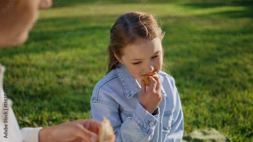 Cute girl eating bun looking on unknown mother resting summer picnic closeup Cute girl eating bun looking on unknown mother resting summer picnic closeup
