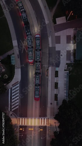 Aerial footage of two streetcars passing each other on a curved road at dusk, surrounded by pedestrian pathways, urban green spaces, illuminated crosswalks, and small buildings.