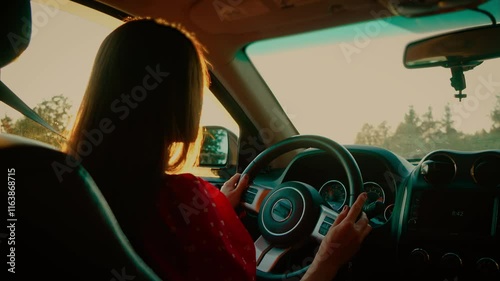 Young woman with straight hair driving car in red dress, holding steering wheel during sunset. Beautiful female focusing on road, evening light illuminating dashboard and interior. Calm lady steering
