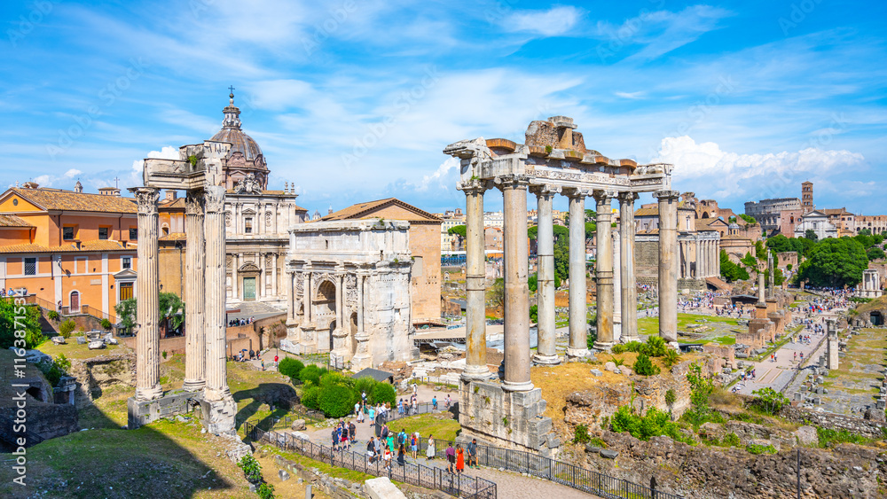 Fototapeta premium Visitors admire the ancient ruins of the Saturn Temple in the Roman Forum, surrounded by historical structures under a clear blue sky. A perfect day for exploring Italy's rich heritage.