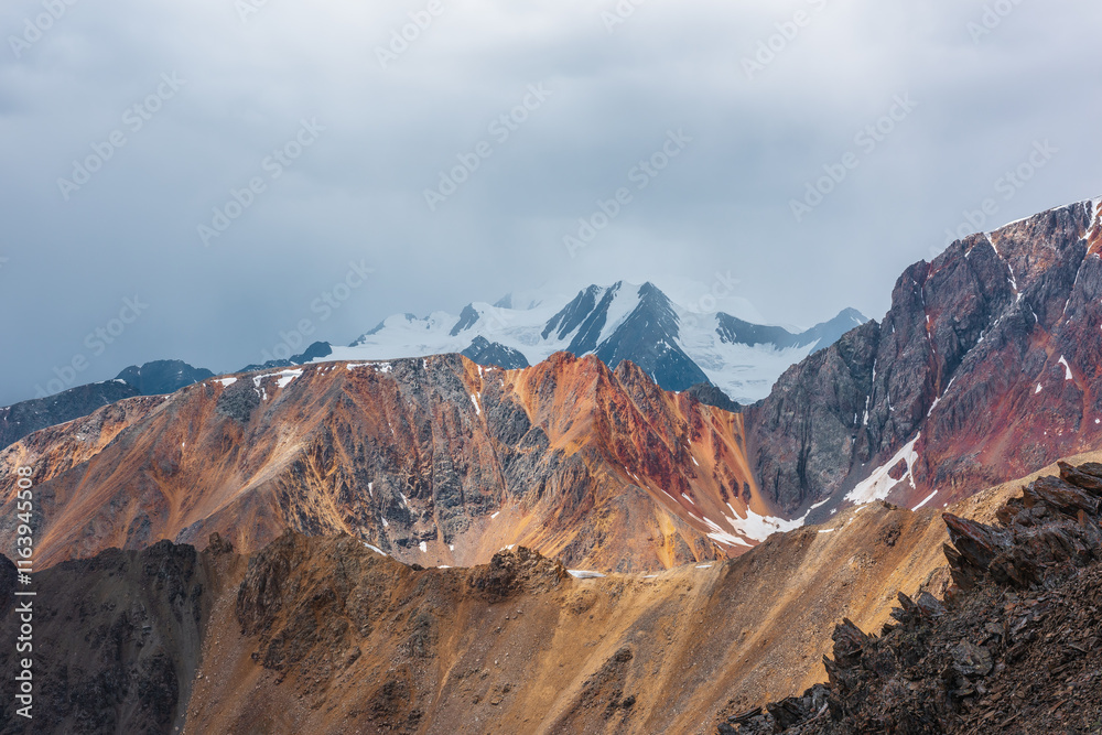 Awesome aerial view to sunlit sharp rocks on ridge of vivid colors and snowy peak in low clouds. Scenic misty landscape with colorful sheer crags against snow-capped mountain top under gray rainy sky. Awesome aerial view to sunlit sharp rocks on ridge of vivid colors and snowy peak in low clouds. Scenic misty landscape with colorful sheer crags against snow-capped mountain top under gray rainy sky.