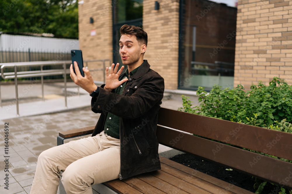 Obraz premium Happy young man in casual clothes having online video chat on smartphone sitting on urban bench near modern office building in city street, waving to screen. Concept of modern communication, lifestyle