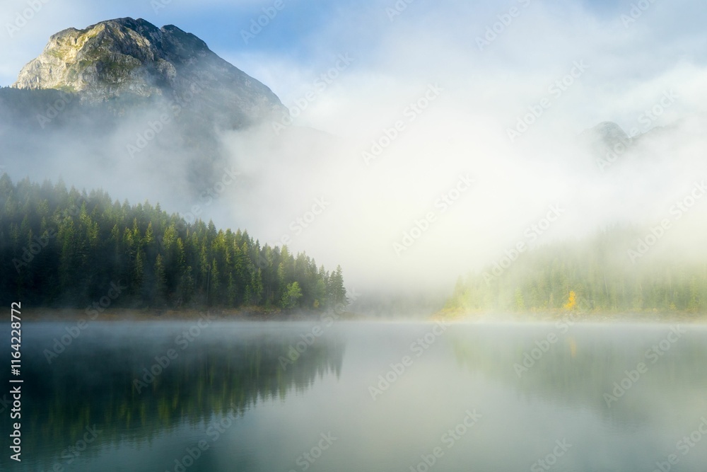 Misty morning over a serene mountain lake with dense forest and a towering peak in the background