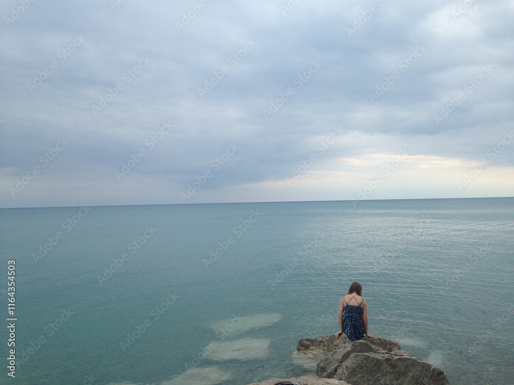 landscape of Lake Huron and a cloudy sky, with a girl in a blue sundress sitting on a rock on the shore landscape of Lake Huron and a cloudy sky, with a girl in a blue sundress sitting on a rock on the shore