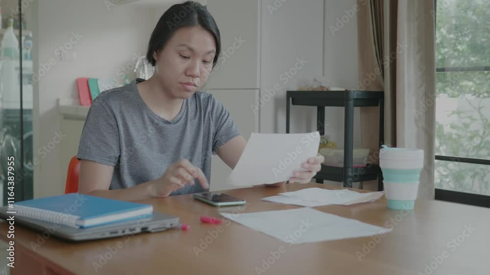 A woman is sitting at a table with a laptop and a cell phone. She is looking at a piece of paper and she is in a serious mood A woman is sitting at a table with a laptop and a cell phone. She is looking at a piece of paper and she is in a serious mood