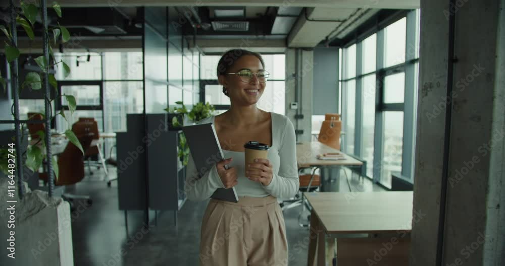 Modern professional woman with glasses holding a coffee cup and laptop walking confidently in a bright office space with panoramic city views and contemporary interior design