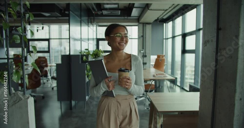 Modern professional woman with glasses holding a coffee cup and laptop walking confidently in a bright office space with panoramic city views and contemporary interior design