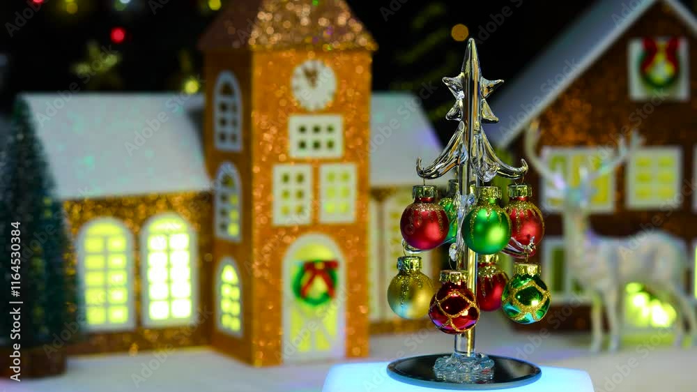 A small glass decorated Christmas tree on a stand with multi-colored lighting rotates against the background of a chapel and a brown house