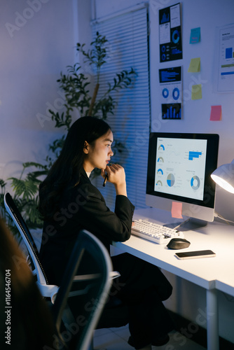 Young businesswoman sitting at her desk in a modern office, working late at night, analyzing charts and graphs on her computer screen, focused on her tasks