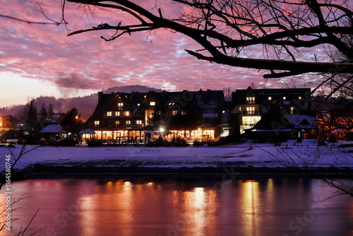 Obraz na płótnie SROMOWCE NIZNE, POLAND - DECEMBER 28, 2024: Beautifully illuminated hotel and restaurant seen at sunset from the opposite bank of the river. w salonie