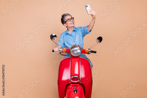 Charming senior woman takes joyful selfie on a vintage red scooter in front of a stylish beige background, embracing modern lifestyle