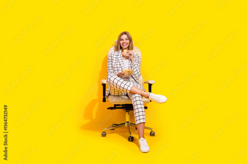 Fototapeta premium Confident young businesswoman in an elegant suit sits on office chair against a yellow background, exuding charm and professionalism.