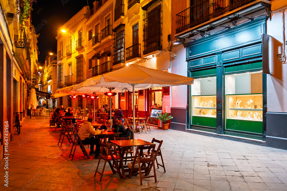 Naklejka premium Sidewalk cafes and shops colorfully illuminated at night in the central Barrio Santa Cruz district of the Andalusian city of Seville, Spain.