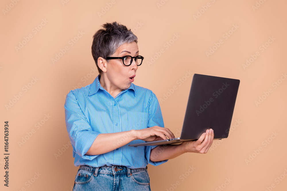 Surprised mature woman holding laptop on beige background in casual striped shirt, showcasing enthusiasm and curiosity Surprised mature woman holding laptop on beige background in casual striped shirt, showcasing enthusiasm and curiosity