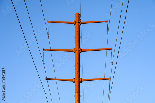 Closeup of upgraded to 230 kV overhead power line distribution system, high-tension power lines on monopole pylon towers, rust orange against a sunny blue sky
