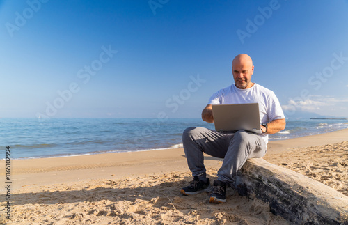 An adult Caucasian man, sitting on a log on the beach, working with a laptop