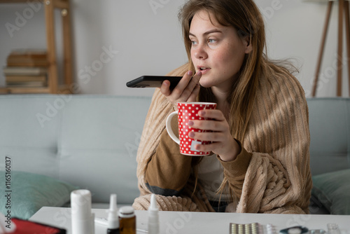 Sickness and virus concept. Woman dictate voice message on phone being sick with flu drinking tea while sitting on sofa suffer from fever