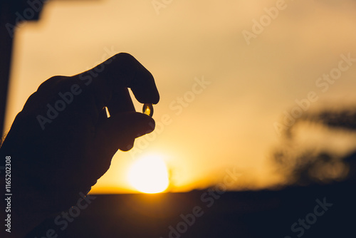 Image of a hand holding a shiny yellow vitamin pill against a sunny orange-golden sky in the morning and evening.