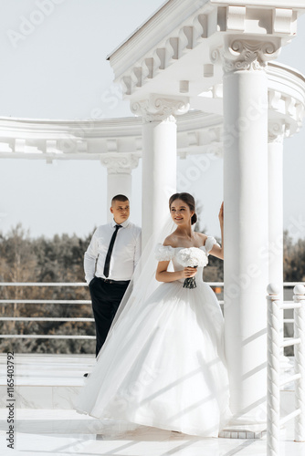 The bride and groom stand against the backdrop of a white architectural structure with columns, reminiscent of a gazebo or pavilion. Trees and a clear sky are visible in the background. photo