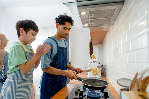 Side view young Asian father show skill of cooking by throwing pan cake up and his son cheer up beside in the kitchen of their house. photo