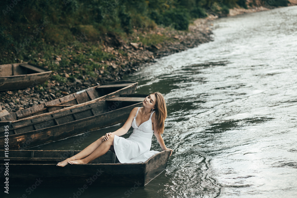 a woman in a white dress sits on a wooden boat that is on the river. She leans back relaxed, resting her hand on the edge of the boat, and looks up. a woman in a white dress sits on a wooden boat that is on the river. She leans back relaxed, resting her hand on the edge of the boat, and looks up.