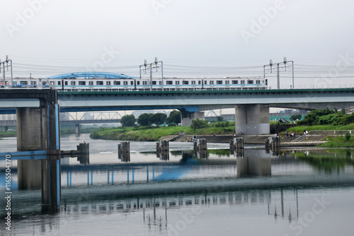 A Shinkansen train crosses a bridge over a calm river, reflecting its structure in the water, surrounded by green banks and distant industrial areas of Tokyo. photo