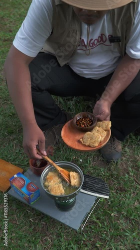 The Bushcraft concept cooks fried tempeh with soy sauce in a dense and green pine forest area. An Asian male solo adventurer is in the process of preparing and cooking in beautiful nature