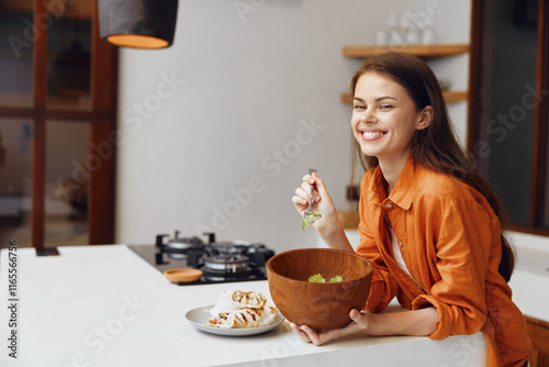 Young woman enjoying a healthy salad in a modern kitchen, smiling and dressed in an orange shirt, emphasizing healthy eating and joyful cooking