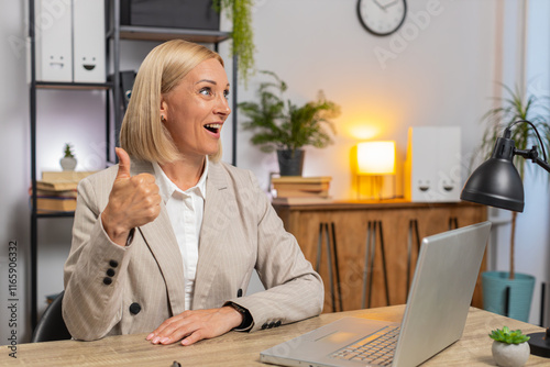 Portrait of smiling mature Caucasian businesswoman freelancer in formal suit showing thumb up, recommending online computer program while sitting in front of laptop netbook at workplace in home office photo