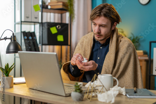 Sick ill young businessman suffering from cold flu or allergy working on laptop at home office. Freelancer bearded man wrapped in blanket blows sneezes wipes snot into napkin. Coronavirus quarantine.