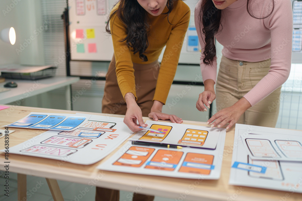 Fototapeta premium Two asian ux designers working together on a new project, pointing at wireframes and discussing user interface improvements for a mobile application