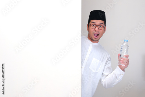 Muslim man peeking out behind a wall showing wow face while holding drinking bottle photo