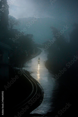 Just after the rain, the evening was dark and bike rider rides his bike on wet road with railway track by the side.